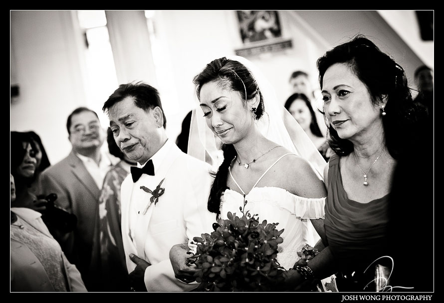 An emotional brides walks down the aisle escorted by her parents. Wedding ceremony at the Holy Ghost Mission Church. Waiakoa, Maui Hi