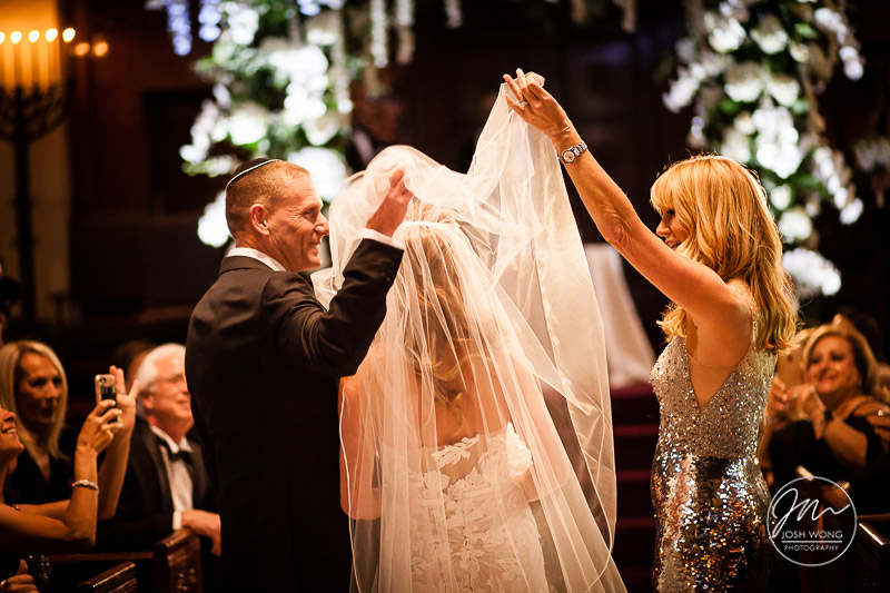 Parents of the bride lift the wedding veil to reveal the bride to the groom after they escort their daughter down the aisle. A Jewish wedding at the Temple of Israel in Lawrence, NY by top New York Wedding Photographers Josh Wong Photography