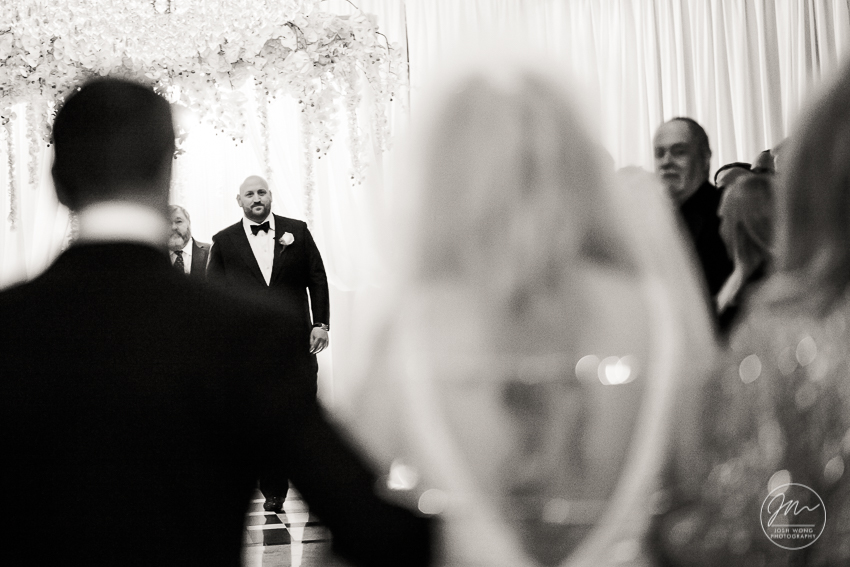 The groom watches intently as the bride walks down the aisle escorted by her parents. Best wedding candid moments at The Venetian in Garfield, NJ. Wedding photos by Josh Wong Photography